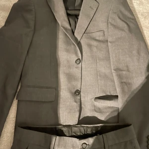 Grå kostym från Lindbergh - Snygg grå kostym från Lindbergh med kavaj och matchande byxor. Kavajen har klassisk design med knappar och fickor. Perfekt för formella tillfällen. Jag är 172 och tycker den sitter lite för stor (ej byxorna då) så kostymen bör passa dig perfekt ifall du är runt 174-179. Använd endast 1 gång