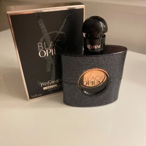 Parfym Black Opium från Yves Saint Laurent - Säljer  Black Opium Eau de Parfum från Yves Saint Laurent 50 ml, med original förpackning. Flaskan är glittrande svart med en rund öppning som visar den gyllene vätskan inuti. Doften är känd för sina djärva och sensuella toner. Perfekt för den som vill ha en elegant och mystisk parfym. Passar perfekt för någon som tycker om att dofta blommor och sött. Endast testad en gång. Säljer p.g.a. inte min smak. Köpt för 979kr på kicks. Hör av dig vid frågor💞
