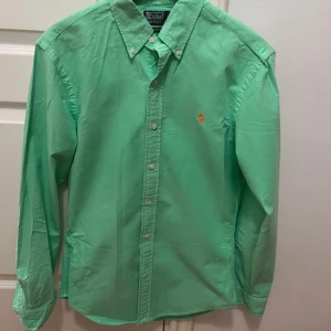 Grön skjorta från Ralph Lauren - Snygg grön skjorta från Ralph Lauren i slim fit. Skjortan har långa ärmar och knappar framtill. Den är tillverkad i bomull och har en broderad logotyp i orange på bröstet. Perfekt för en stilren look!