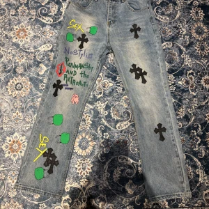 Chrome hearts x Levi’s Jeans - Säljer ett par unika chrome hearts x Levi's jeans med coola korsdetaljer och färgglada tryck. Jeansen har en straight passform och är i en klassisk blå denim. Perfekta för dig som vill sticka ut med en edgy stil.