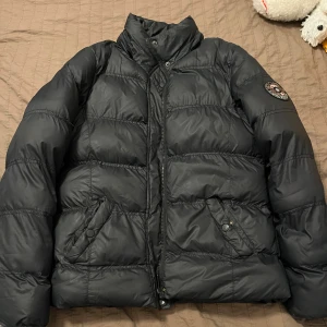 Svart dunjacka med emblem - Snygg svart dunjacka med hög krage och emblem på ärmen. Jackan har dragkedja och knappar framtill samt praktiska fickor med knäppning. Perfekt för kyliga dagar. Jackan är specifikt en vårjacka.💕