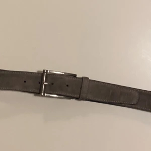 Grått mockabälte från Tiger of Sweden - Snyggt grått bälte i äkta skinn från Tiger of Sweden. Bältet har en stilren design med en silverfärgad spänne. Perfekt för att ge en elegant touch till din outfit.