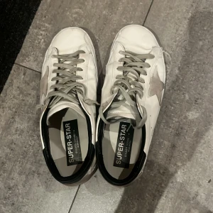 Super-Star sneakers - Säljer ett par vita Golden Goose Super-Star sneakers med grå detaljer och snörning. Skorna har en klassisk design med en stjärna på sidan och svart häl. Perfekta för en avslappnad stil. Köpta i andra hand. Skicket på skorna förklarar priset 