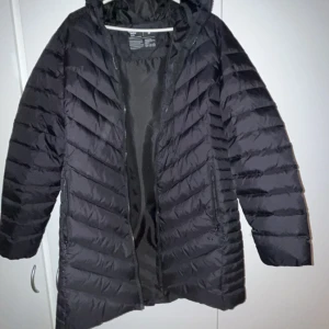 Svart dunjacka från Neomondo - Säljer en svart dunjacka från Neomondo. Jackan har en lång design med dragkedja och är perfekt för kyliga dagar. Den är quiltad och har långa ärmar, vilket gör den både stilren och funktionell. Både vattentät och vindtät