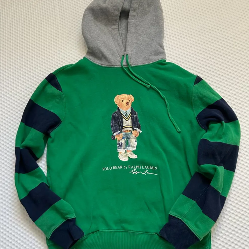 Säljer en supercool grön hoodie från Ralph Lauren med ett gulligt björnmotiv på framsidan. Den har en grå huva och mörkblå ränder på ärmarna. Perfekt för en avslappnad stil och passar bra till både jeans och joggers. Den är i bra skick och redo för nya äventyr! 🐻💚. Tröjor & Koftor.