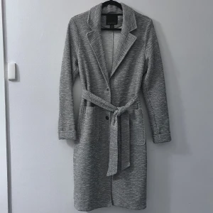 Grå kappa - Säljer en snygg grå kappa i storlek L-XL. Den har en klassisk design med knappar och ett bälte i midjan för en elegant look. Perfekt för höst och vår, med långa ärmar och en stilren krage. ❤️