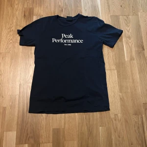 Svart t-shirt från Peak Performance - Säljer en stilren svart t-shirt från Peak Performance. Den har en klassisk design med vit text på framsidan och är perfekt för vardagsbruk. T-shirten är i bomull och har korta ärmar. Perfekt för en avslappnad look!