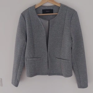 Grå kavaj från Vero Moda - Säljer en stilren grå kavaj från Vero Moda. Den har ett snyggt quiltat mönster och är perfekt för både vardag och fest. Kavajen är långärmad och har en modern, minimalistisk design utan krage. Perfekt för höst och vår!