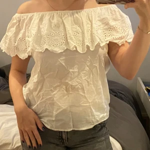 Vit off-shoulder topp med spetsdetaljer - Säljer en superfin vit off-shoulder topp med spetsdetaljer. Den har en luftig och bekväm passform, perfekt för sommardagar. Toppen är ärmlös och har en vacker volang runt axlarna.