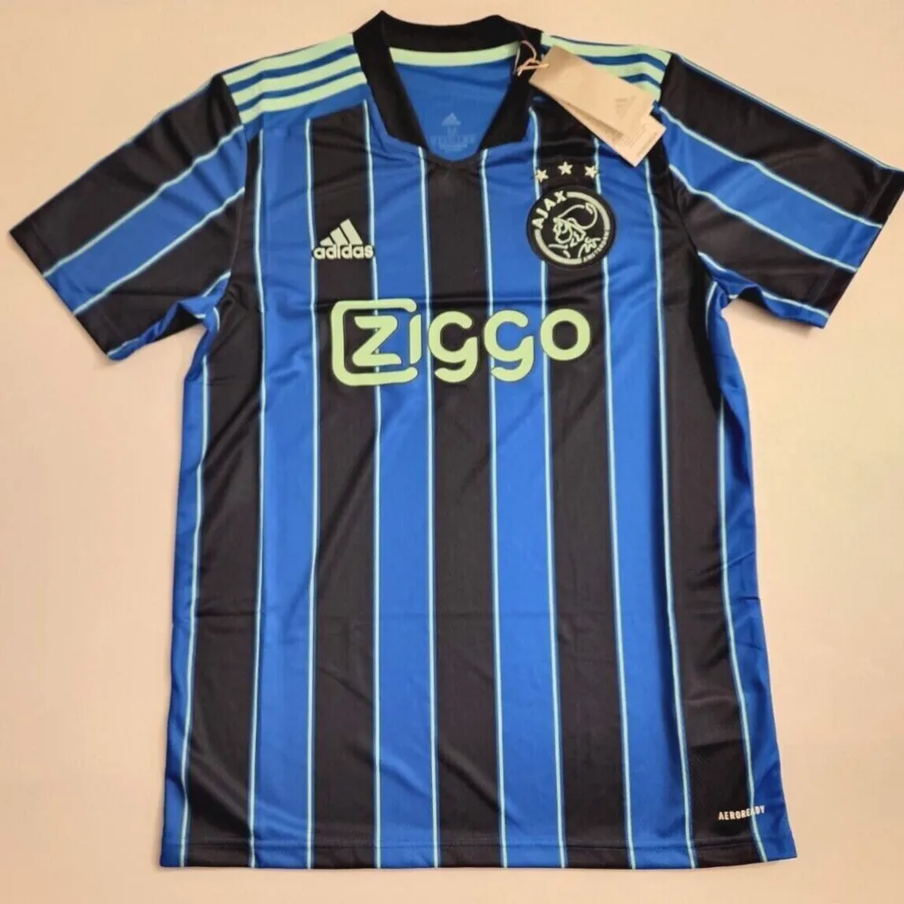 Säljer en snygg fotbollströja från Ajax i blått och svart med randigt mönster. Tröjan har korta ärmar och en klassisk design med klubbens logga och Ziggo som sponsor på framsidan. Perfekt för fotbollsfans!. T-shirts.