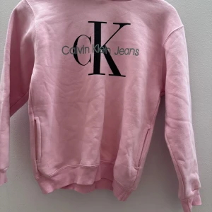 Rosa hoodie från Calvin Klein - Säljer en snygg rosa hoodie från Calvin Klein Jeans med deras ikoniska logga på framsidan. Tröjan har långa ärmar och en bekväm huva. Perfekt för en avslappnad stil. är i storlek 12 