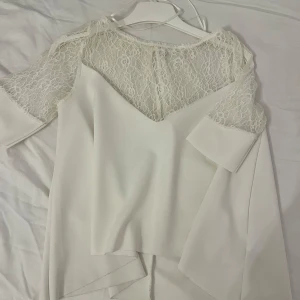 Vit blus med spetsdetaljer - Elegant vit blus med spetsdetaljer på axlar och ärmar. Blusen har en v-ringad design och trekvartsärmar, vilket ger en stilren och feminin look. Perfekt för en chic och sofistikerad stil.