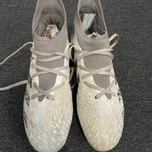 Adidas predator fotbollsskor med grått mönster - Snygga Adidas fotbollsskor med ett unikt grått mönster och tre svarta ränder på sidan. Skorna har snörning och en hög ankelkrage för extra stöd. Sulan är utrustad med röda och gröna dobbar för optimalt grepp på planen. Det är skruv dobbar också 