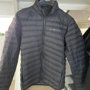 Svart dunjacka från Sail Racing - Snygg svart dunjacka från Sail Racing med deras unika Link Technology. Jackan har en dragkedja framtill och är designad för att ge optimal värme och rörelsefrihet. Perfekt för kyligare dagar.