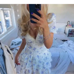 Blommig klänning med volanger - Charmig vit klänning med blått blommönster och volanger. Den har en v-ringad design och korta ärmar, perfekt för sommarens äventyr. Klänningen har en feminin och lekfull stil som passar perfekt för soliga dagar.