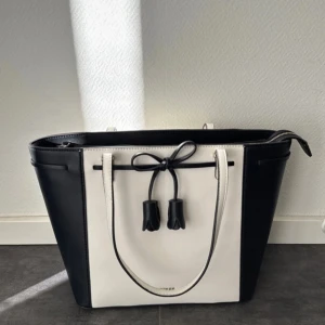 Svart och vit axelväska från Guess - Snygg svart och vit axelväska från Guess med stilren design. Väskan har en dekorativ knytning framtill och en praktisk dragkedja på baksidan. Perfekt för att bära med sig det viktigaste med stil.