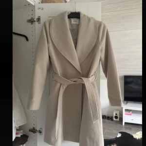 Beige kappa från JDY - Snygg beige kappa från JDY med knytbälte i midjan. Den har en stilren design med långa ärmar och knappar framtill. Perfekt för kyligare dagar.