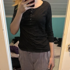 Svart långärmad topp med knappar - Säljer en svart långärmad topp med knappar framtill. Perfekt för en stilren och enkel look. Toppen har en bekväm passform och trekvartsärmar.