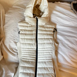 Créme färgad dunväst från Zara - Snygg vit/créme färgad dunväst från Zara med huva och dragkedja framtill. Perfekt för kyliga dagar när du vill hålla dig varm och stilren. Aldrig använd, den har bara hängt i garderoben. Dock ingen lapp kvar.💓