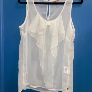 Vit ärmlös blus med rosettdetalj från Only - Säljer en stilren vit ärmlös blus från Only. Blusen har en elegant volangdetalj som liknar en rosett framtill och är tillverkad i ett lätt och luftigt material. Använd fåtal gången, inga skador, storlek 36. Pris kan diskuteras!