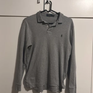 Grå pikétröja från Ralph Lauren - Säljer en klassisk grå pikétröja från Ralph Lauren med lång ärm och den ikoniska loggan broderad på bröstet. Perfekt för en stilren och avslappnad look. Tröjan har en krage och knappar framtill. Den är i bra skick och inga defekter. Skulle passa nån som är mellan 170+