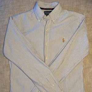 Ralph Lauren skjorta - Snygg blå- och vitrandig skjorta från Ralph Lauren med klassisk krage och broderad logga på bröstet. Skjortan har långa ärmar och knappar framtill. Perfekt för en stilren look.