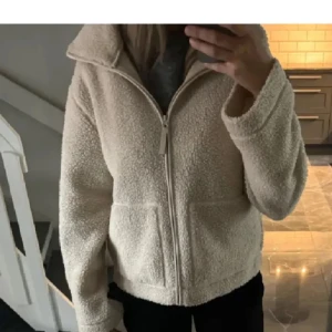 Beige teddyjacka med dragkedja - Mysig beige teddyjacka med dragkedja framtill och hög krage. Jackan har långa ärmar och två stora fickor fram. Perfekt för kyliga dagar när du vill hålla dig varm och bekväm.