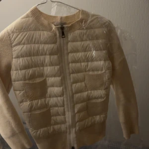 Beige quiltad jacka från Moncler cardigan  - Säljer en snygg beige jacka från Moncler med quiltad design och dragkedja framtill. Jackan har långa ärmar och stickade detaljer vid fickorna. Perfekt för kyligare dagar och ger en stilren look. Jackan är storlek s men sitter som xs har en skada som syns på bild jackan e äkta å e köpt på vestiaire 
