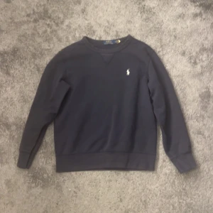 Mörkblå tröja från Polo Ralph Lauren - Säljer en stilren mörkblå tröja från Polo Ralph Lauren med det klassiska broderade logotypen på bröstet. Tröjan har långa ärmar och en rund halsringning med ribbade muddar vid ärmslut och nederkant. Perfekt för en avslappnad look.