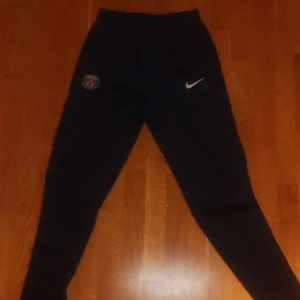 Svarta träningsbyxor från Nike med PSG-logga - Snygga svarta träningsbyxor från Nike med PSG-logga på ena benet. Byxorna har en elastisk midja och en smal passform som ger en sportig look. Perfekta för fotbollsträningen eller gymmet.