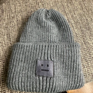 Grå stickad mössa med broderad patch - Säljer en stilren grå stickad mössa med en broderad patch framtill. Mössan har en ribbad design som ger en skön och flexibel passform. Perfekt för kyliga dagar.