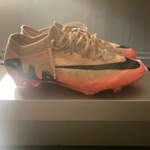 Nike Mercurial fotbollsskor - Snygga Nike Mercurial fotbollsskor i beige med svarta och orange detaljer. Skorna har snörning och en modern design som passar perfekt för fotbollsspelare. De är lätta och designade för att ge bra grepp på planen.
