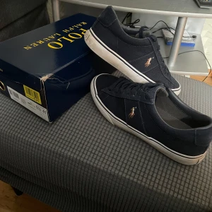  Sneakers från Polo Ralph Lauren - Snygga marinblå sneakers från Polo Ralph Lauren med klassisk design och broderad logotyp på sidorna. Skorna har en vit sula och snörning framtill. Perfekta för en stilren look. Knappt använda pris kan diskuteras!
