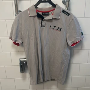 Grå pikétröja från Sail Racing - Säljer en grå pikétröja från Sail Racing i storlek L. Tröjan har korta ärmar och detaljer i rött och svart vid kragen. Aldrig använd stilren. Försöker frakta samma dag 