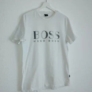 Vit t-shirt från Hugo Boss - Säljer en stilren vit t-shirt från Hugo Boss med tryck på framsidan. T-shirten har en normal passform och är kortärmad, perfekt för en avslappnad look. Den är tillverkad i ett material som erbjuder solskydd.