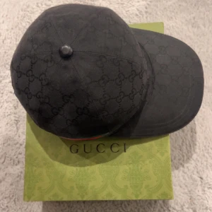 Svart keps från Gucci - Snygg svart keps från Gucci med deras ikoniska mönster. Kepsen har en röd och grön randdetalj på sidan. Perfekt för att ge din outfit en lyxig touch.