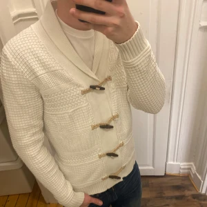 Vit stickad cardigan  - Säljer en stilren vit stickad kofta med unika träknappar och snörning framtill. Koftan har ett snyggt mönster och ribbade muddar vid ärmslut och nederkant. Perfekt för kyliga dagar! Modellen är ca 182, tveka inte att kontakta vid eventuella frågor 😀