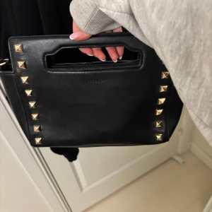 Svart handväska med nitar från Picard - Snygg svart handväska från Picard med coola guldfärgade nitar längs kanterna. Väskan har ett praktiskt handtag och en dragkedja för säker förvaring. Väskan har ett tillkommande axelband.