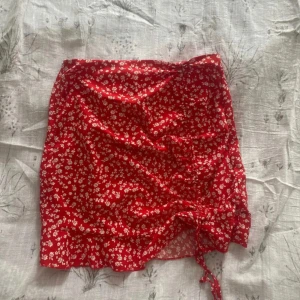 Röd blommig kjol från Shein - Snygg röd blommig kjol från Shein i storlek S. Kjolen har ett fint blommönster och en knytning framtill som ger en extra detalj. Perfekt för sommarens varma dagar!