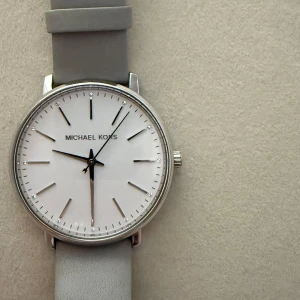 Grå klocka från Michael Kors - Stilren klocka från Michael Kors med en grå skinnrem och en vit urtavla. Klockan har en silverfärgad boett och minimalistiska markeringar. Perfekt för den som gillar en enkel och elegant stil 🩶