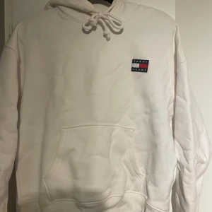 Vit hoodie från Tommy Jeans - Säljer en stilren vit hoodie från Tommy Jeans. Den har en klassisk design med en liten logotyp på bröstet och en praktisk känguruficka framtill. Perfekt för en avslappnad look.