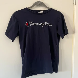 Mörkblå t-shirt från Champion - Säljer en mörkblå t-shirt från Champion med deras ikoniska logga broderad på bröstet. T-shirten är kortärmad och tillverkad i mjuk bomull. Perfekt för en avslappnad stil. Den är i barn storlek 