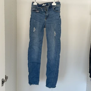 Blå jeans med slitningar - Snygga blå jeans med slitningar på framsidan. De har en klassisk femficksdesign och en dragkedja. Perfekta för en avslappnad stil.