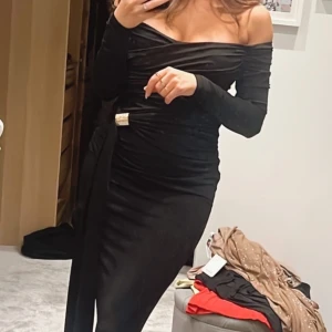 Svart långklänning med offshoulder-design - Elegant svart långklänning med offshoulder-design och långa ärmar från Labelnue. Klänningen har en smickrande drapering och en dekorativ detalj i midjan. Perfekt för en stilfull kväll. Stretchig och skönt material. Endast använd en gång så i fint skick!