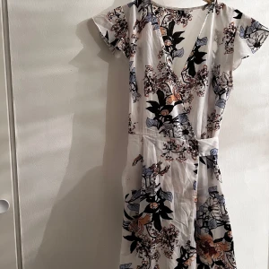 Blommig långklänning med slits - Säljer en vacker långklänning med blommigt mönster i svart, blått och beige. Klänningen har en v-ringad design och korta ärmar. Den har en elegant slits framtill som ger en fin touch. Perfekt för sommarens alla tillfällen!