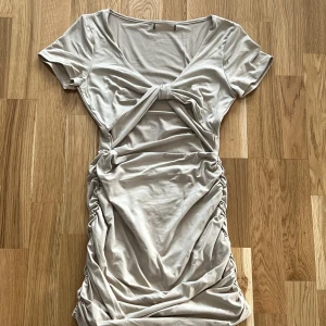 Beige klänning med knytdetalj - Trendig beige klänning med knytdetalj framtill och rynkade sidor. Den har korta ärmar och en v-ringad design som ger en stilren look. Perfekt för att skapa en chic outfit.