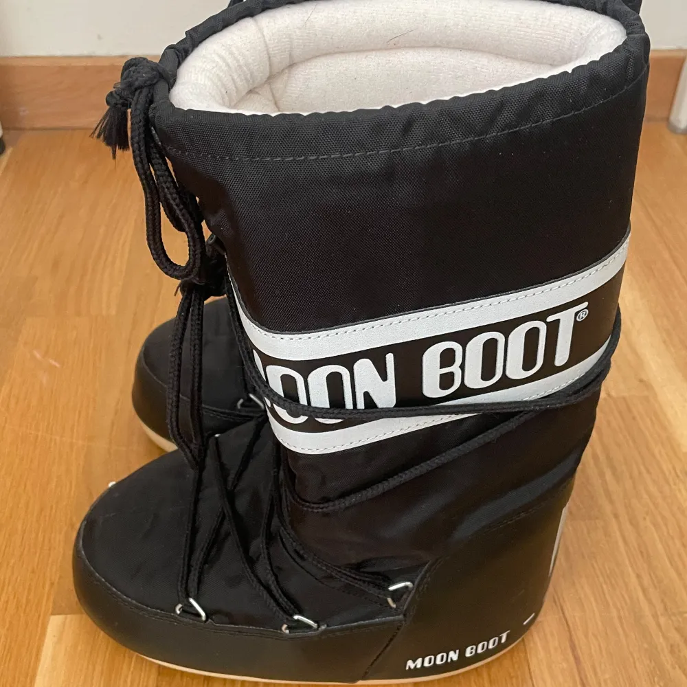 Säljer ett par svarta Moon Boots med vit text och snörning. Perfekta för vintern med sin varma och vadderade design. De har en hög modell och är kända för sin unika stil och komfort.. Kengät.