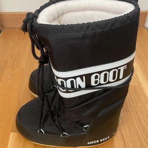 Svarta Moon Boots - Säljer ett par svarta Moon Boots med vit text och snörning. Perfekta för vintern med sin varma och vadderade design. De har en hög modell och är kända för sin unika stil och komfort.
