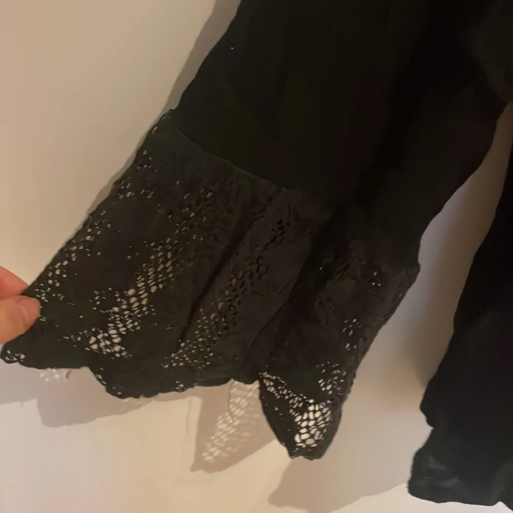 Säljer en elegant svart blus med vackra spetsdetaljer vid halsringningen och ärmsluten. Blusen har långa ärmar och en lös passform, perfekt för en stilren look.. Yläosat.
