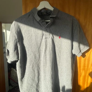 Grå pikétröja från Ralph Lauren - Säljer en klassisk grå pikétröja från Ralph Lauren med den ikoniska röda broderade loggan på bröstet. Tröjan har korta ärmar och en stilren krage med knappar. Perfekt för en avslappnad men ändå stilfull look. Pris kan diskuteras